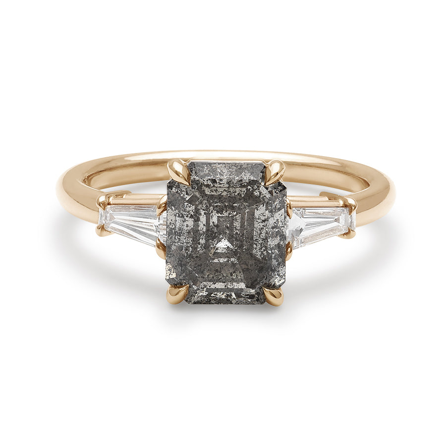 A three stone engagement ring with an emerald shaped grey diamond center stone and two white diamond tapered baguettes on either side set in yellow gold.