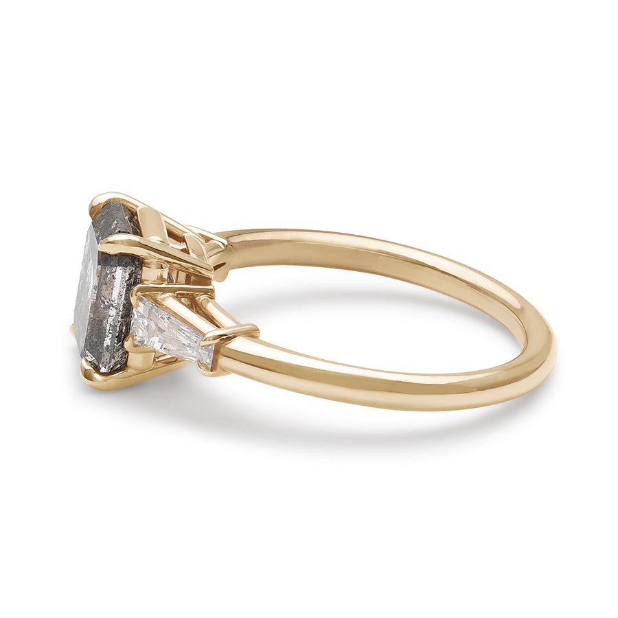 Angled view of a three stone engagement ring with an emerald shaped grey diamond center stone and two white diamond tapered baguettes on either side set in yellow gold.
