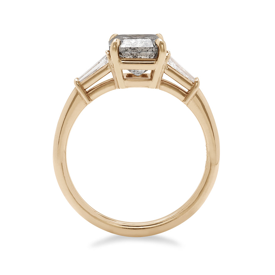 Side view of a three stone engagement ring with an emerald shaped grey diamond center stone and two white diamond tapered baguettes on either side set in yellow gold.