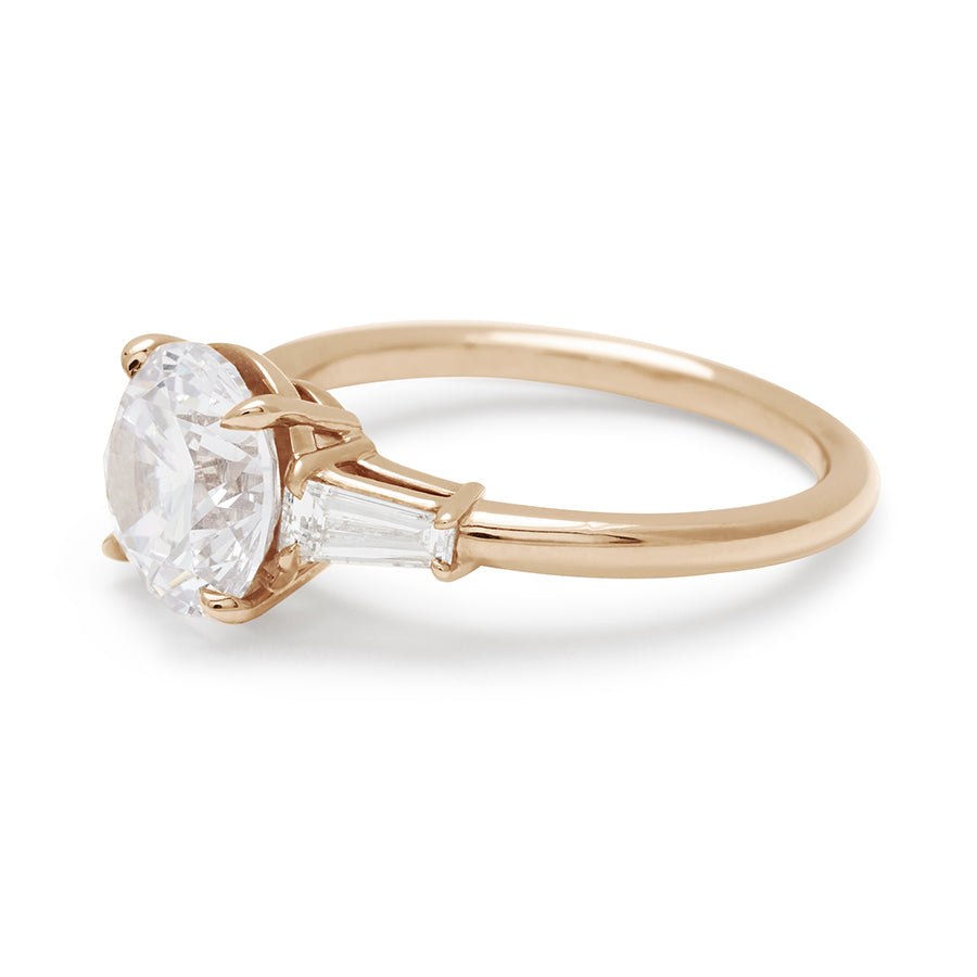 Angled view of a three stone engagement ring with a round white diamond center stone and two white diamond tapered baguettes on either side set in 14k gold.