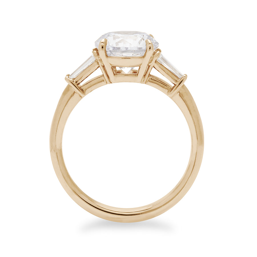 Side view of a three stone engagement ring with a round white diamond center stone and two white diamond tapered baguettes on either side set in 14k gold.