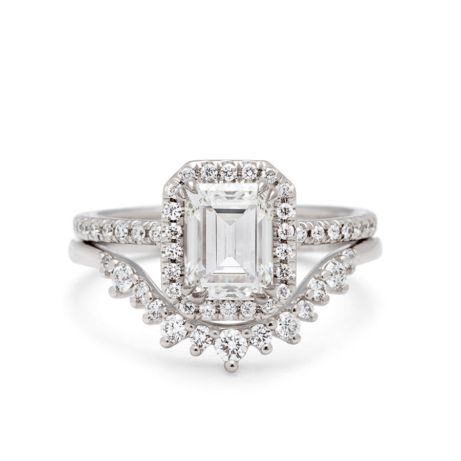 A solitaire engagement ring with an emerald cut and emerald shape white diamond center stone surrounded by a white diamond pave halo with white diamond pave around the band set in white gold set above a 13 white diamond curved band set in white gold.