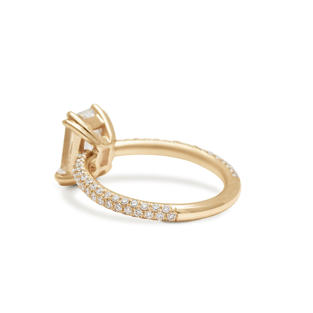 Angled view of a solitaire emerald shape and emerald cut white diamond ring with double claw prongs with three rows of white diamond pave on the band set in yellow gold.