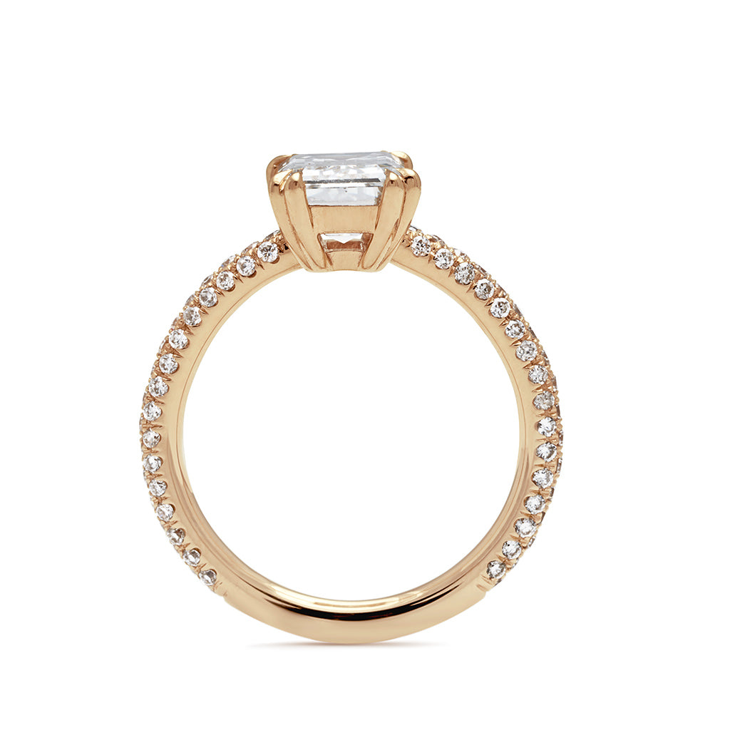 Side view of a solitaire emerald shape and emerald cut white diamond ring with double claw prongs with three rows of white diamond pave on the band set in yellow gold.