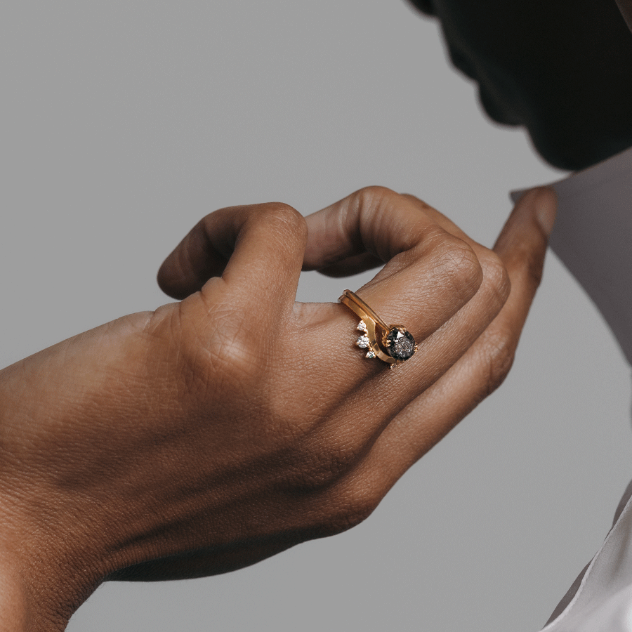 A solid gold band with asymetrical white diamond accent stones set in yellow gold stacked beneath a solitaire east/west grey diamond ring on a dark-complexion ring finger.