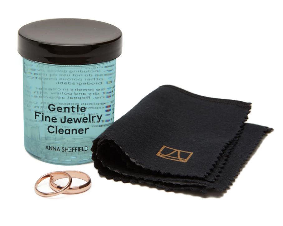 "Anna Sheffield Gentle Fine Jewelry Cleaner" in a small jar with blue liquid with a jewelry cloth next to it and two solid gold bands.