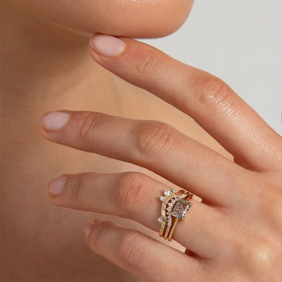 An east/west solitaire ring with an emerald shape black rutilated quartz stone set in yellow gold set below an 11 white diamond curve band and an asymetrical white diamond yellow gold band. On a lighter-complextion hand that is lightly touching their face., White Gold, Rose Gold, Yellow Gold