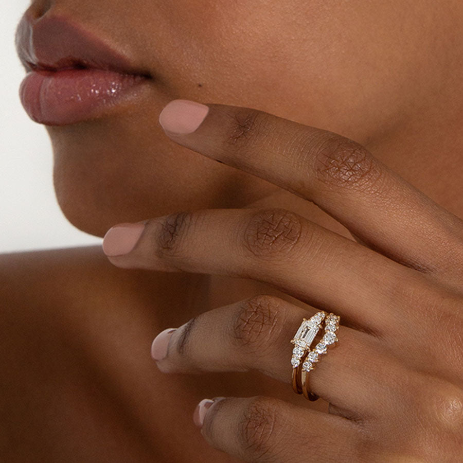 A ring with an emerald cut and shaped white diamond center stone with two round white diamonds on either side set in yellow gold above a 7 round white diamond curve band on a darker complexion model., White Gold, Rose Gold, Yellow Gold