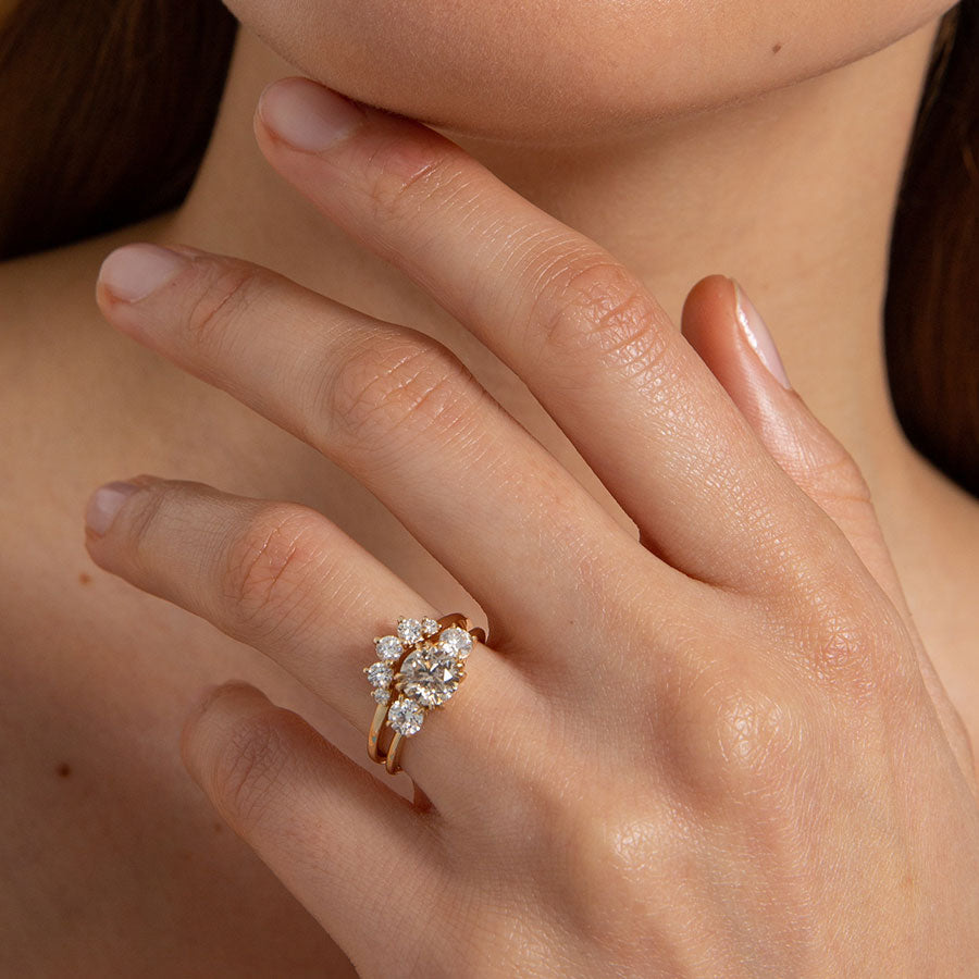 Female Hand by Chin wearing A curved 5 white diamond band set in yellow gold above a three stone ring with a champagne diamond center stone on a light-complexion hand.