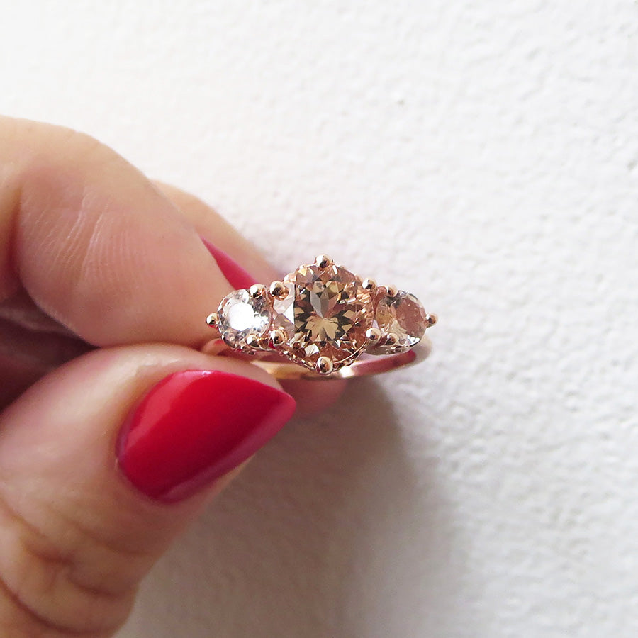 A three stone engagement ring with a round shape pink morganite center stone with two pink morganite round side stones set in rose gold being held by a light-complexion hand with red nails.