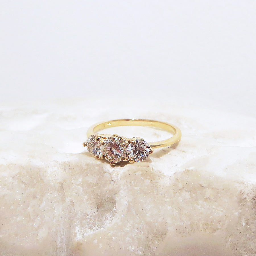 A three stone engagement ring with a round shape champagne diamond center stone with two white diamond round side stones set in yellow gold on a white stone.