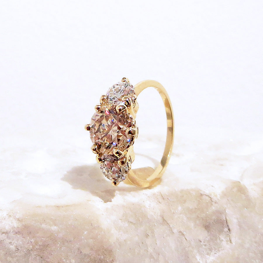 Angled view of a three stone engagement ring with a round shape champagne diamond center stone with two white diamond round side stones set in yellow gold on a pink stone.