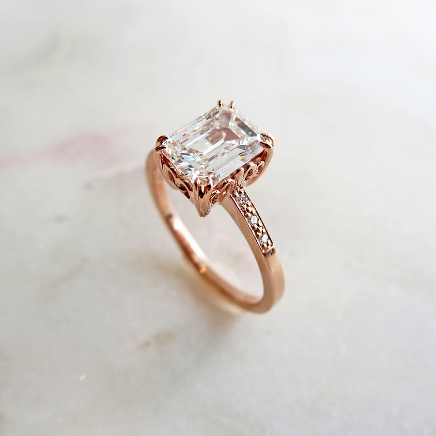 Angled view of a solitaire ring with an emerald shape and cut champagne diamond with white diamond pave on either side set in rose gold on a pink background.