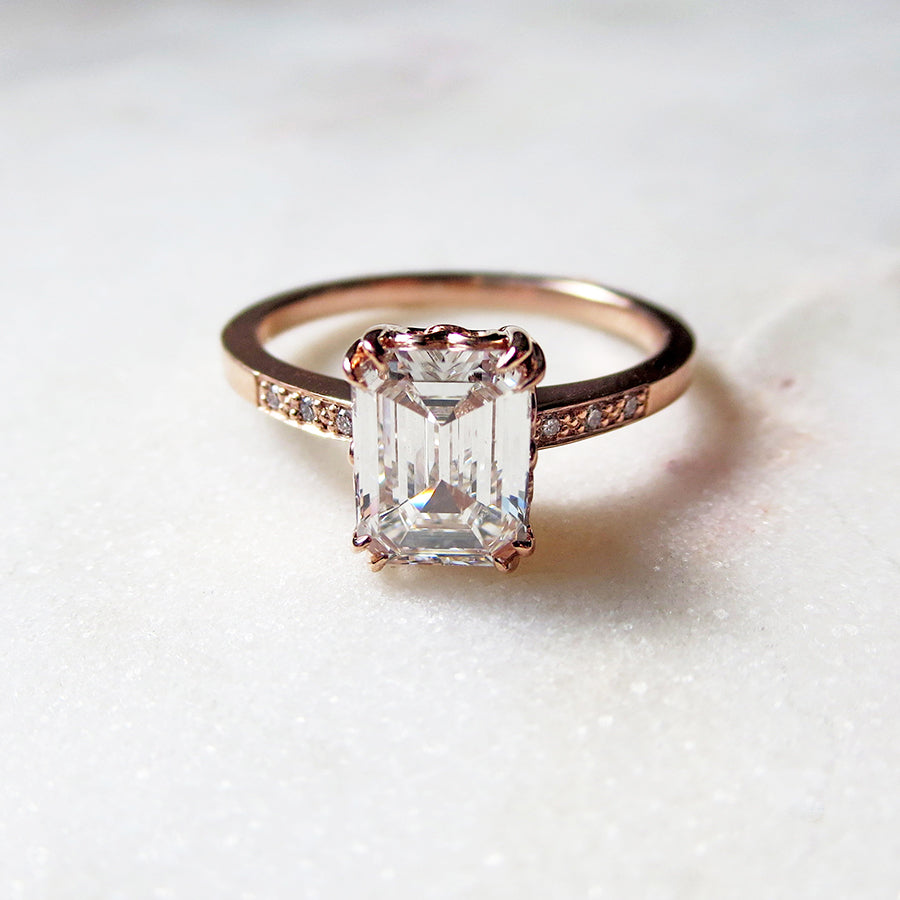A solitaire ring with an emerald shape and cut champagne diamond with white diamond pave on either side set in rose gold on a pink background.