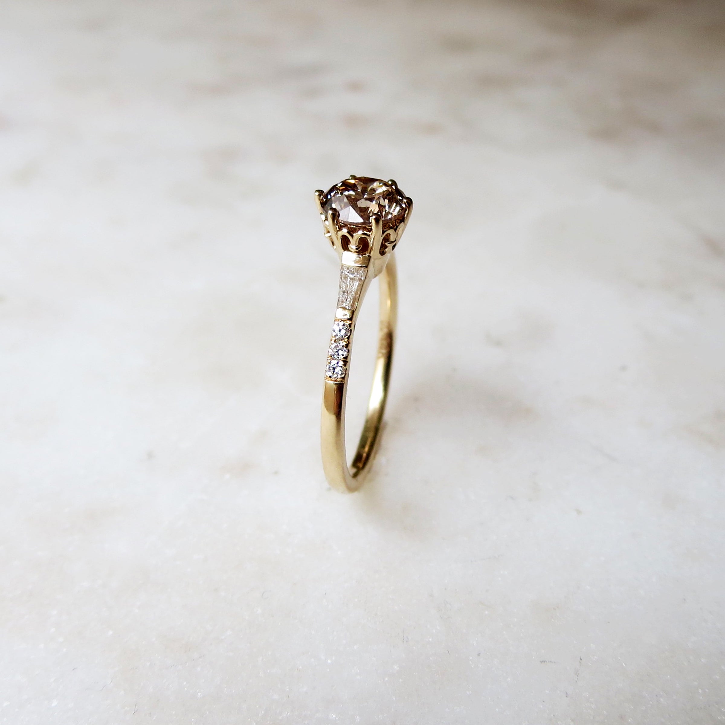 Side view of a solitaire ring with a round champagne diamond with tapered baguette accent stones and white diamond pave on either side set in yellow gold.