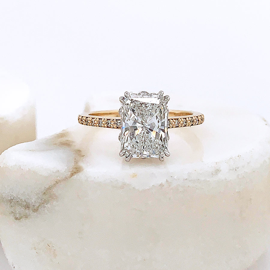 A solitaire emerald cut and shaped white diamond ring with double claw prongs set in white gold basket with white diamond pave on the band set in yellow gold on a grey stone.
