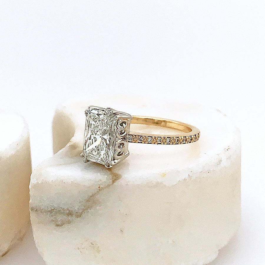 Side view of a solitaire emerald cut and shaped white diamond ring with double claw prongs set in white gold basket with white diamond pave on the band set in yellow gold on a grey stone.