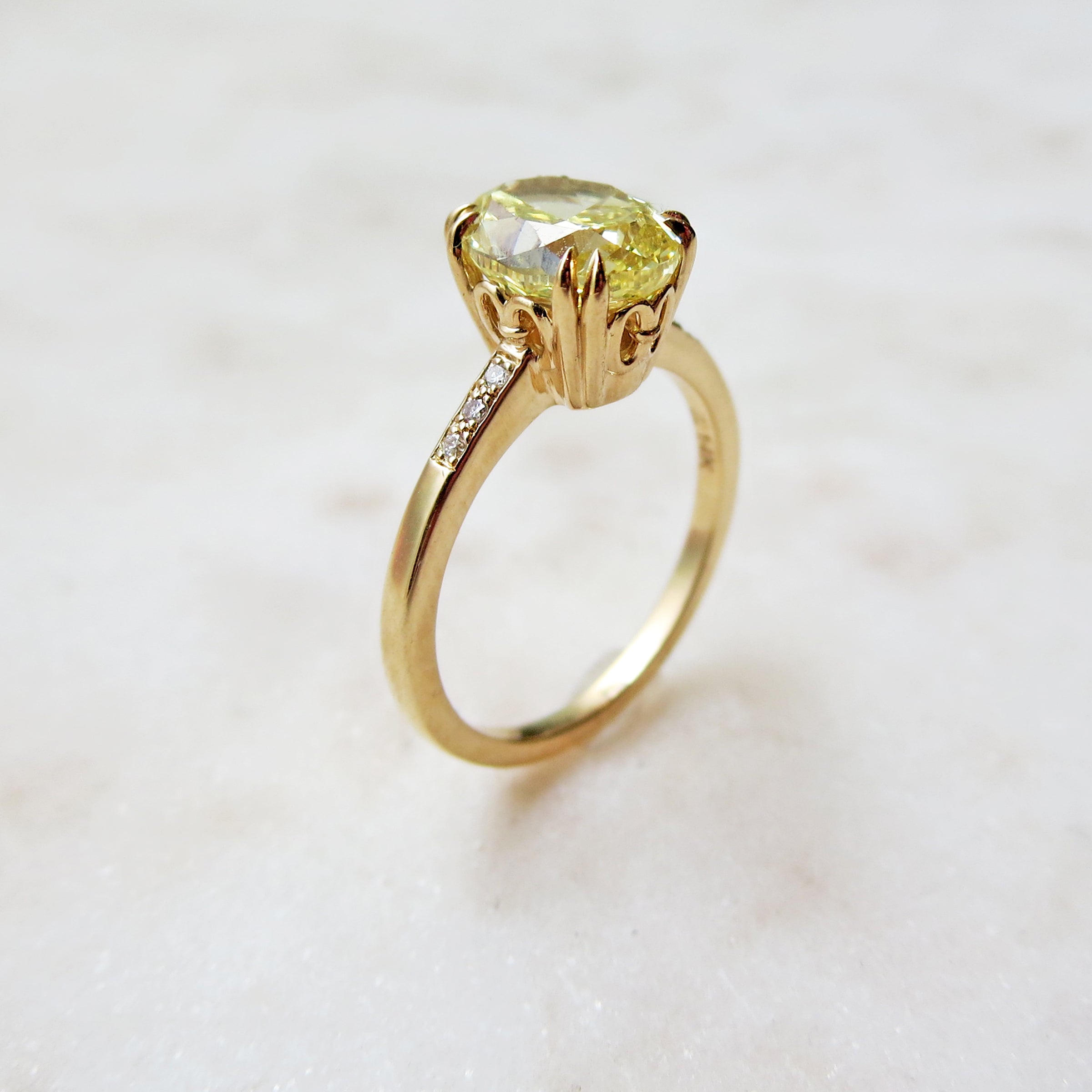 Side view of a solitaire ring with a cushion cut yellow diamond with white diamond pave on either side with double claw prongs set in yellow gold on a grey background.
