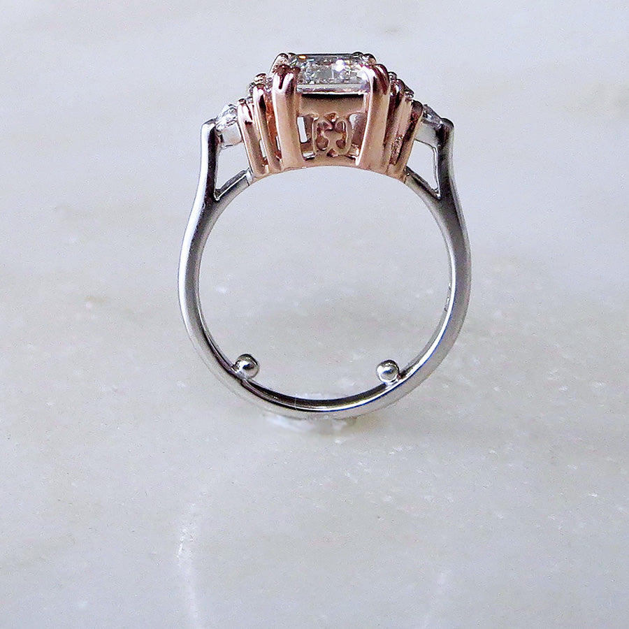 Side view of a ring with an emerald cut and shaped white diamond center stone with one baguette white diamond and one white diamond trillion on either side with rose gold claw prongs set in white gold on a grey background.