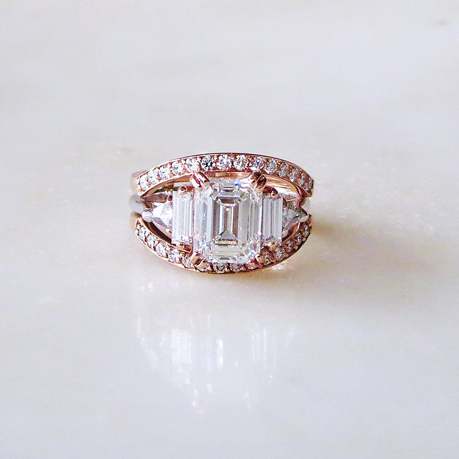 A ring with an emerald cut and shaped white diamond center stone with one baguette white diamond and one white diamond trillion on either side with rose gold claw prongs set in white gold on a grey background stacked between two white diamond dusted curve bands.