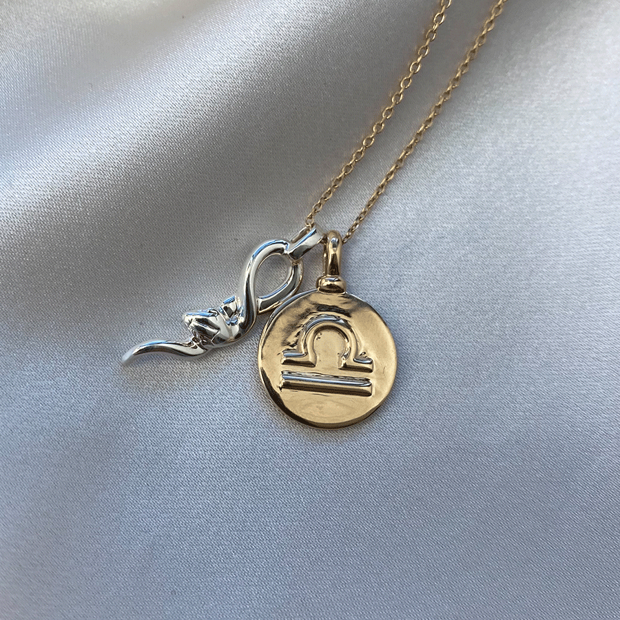 A 14mm charm with the Libra zodiac sign set in yellow gold on a yellow gold chain with a medium serpent charm set in sterling silver next to it on grey fabric.