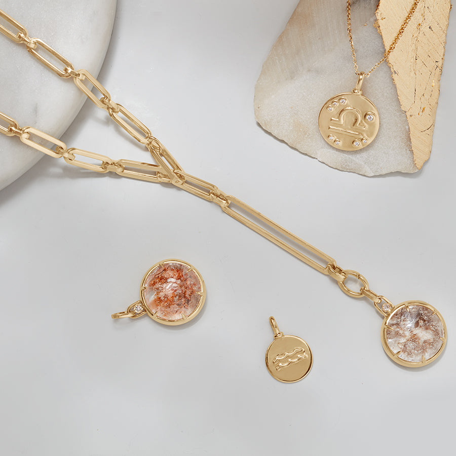 A quartz charm with a desert like scene in a bezel setting with claw prongs set in yellow gold with white diamond accent stone on a mixed link yellow gold chain on a grey background with zodiac charms around it.