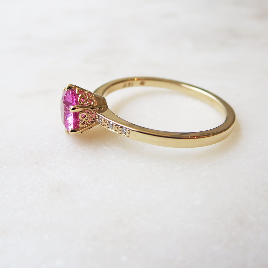 Side view of a solitaire ring with a round pink sapphire stone with white diamond pave on either side set in yellow gold on a grey background.