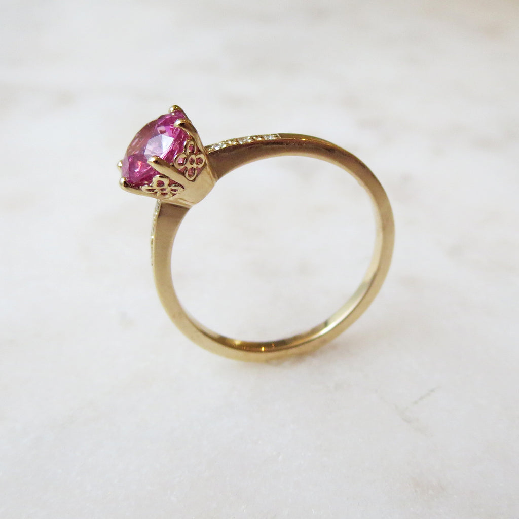 Angled view of a solitaire ring with a round pink sapphire stone with white diamond pave on either side set in yellow gold on a grey background.