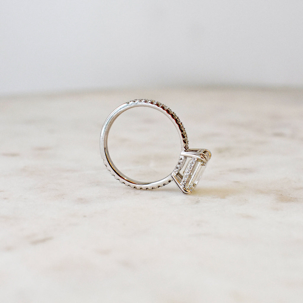 Angled view of a solitaire east/west emerald shape white diamond ring with double claw prongs with white diamond pave on the band set in white gold on a grey background.