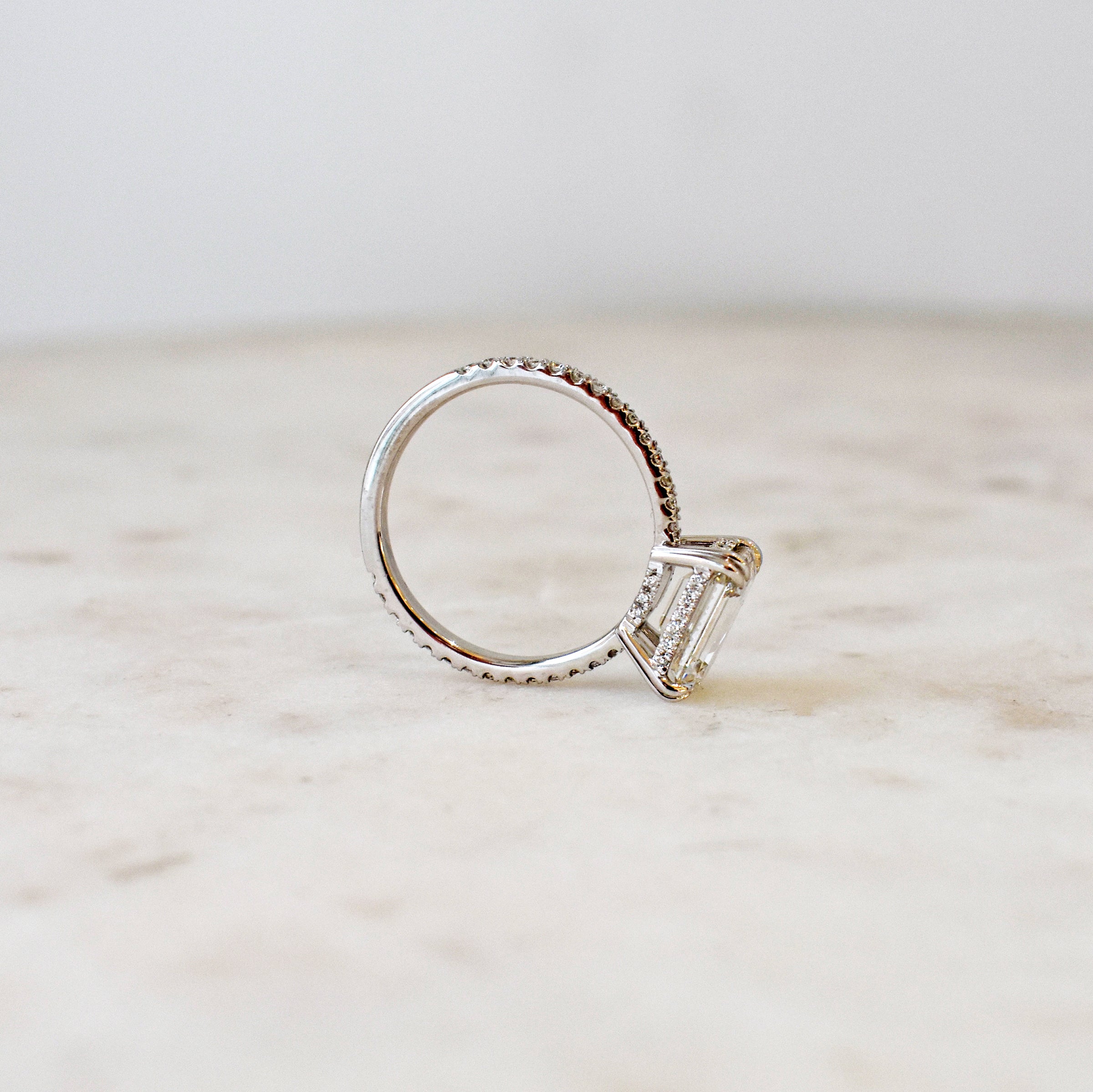 Angled view of a solitaire east/west emerald shape white diamond ring with double claw prongs with white diamond pave on the band set in white gold on a grey background.