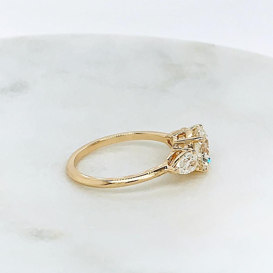 Side view of a three stone engagement ring with a round shape white diamond center stone with two white diamond pear shaped side stones set in yellow gold on a grey background.