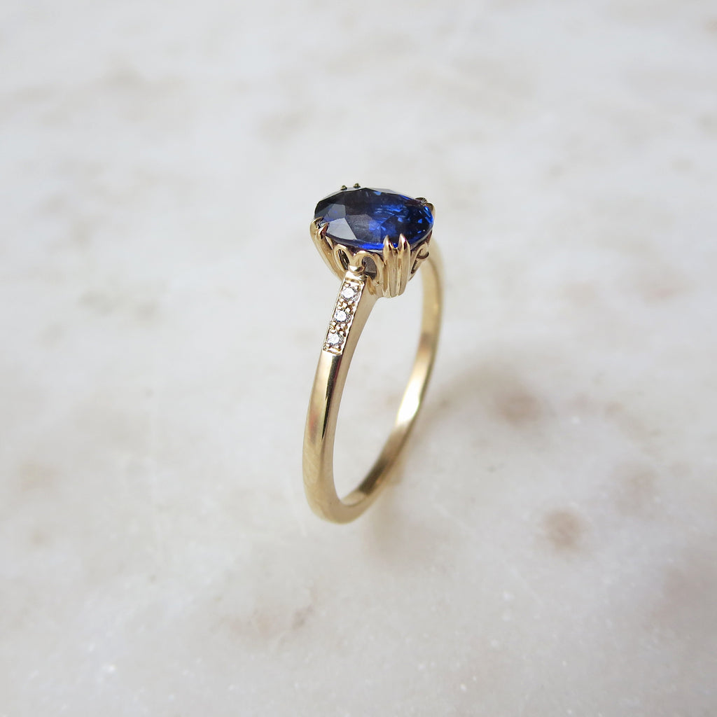 Angled view of a solitaire ring with an oval blue sapphire stone with white diamond pave on either side set in yellow gold on a grey background.