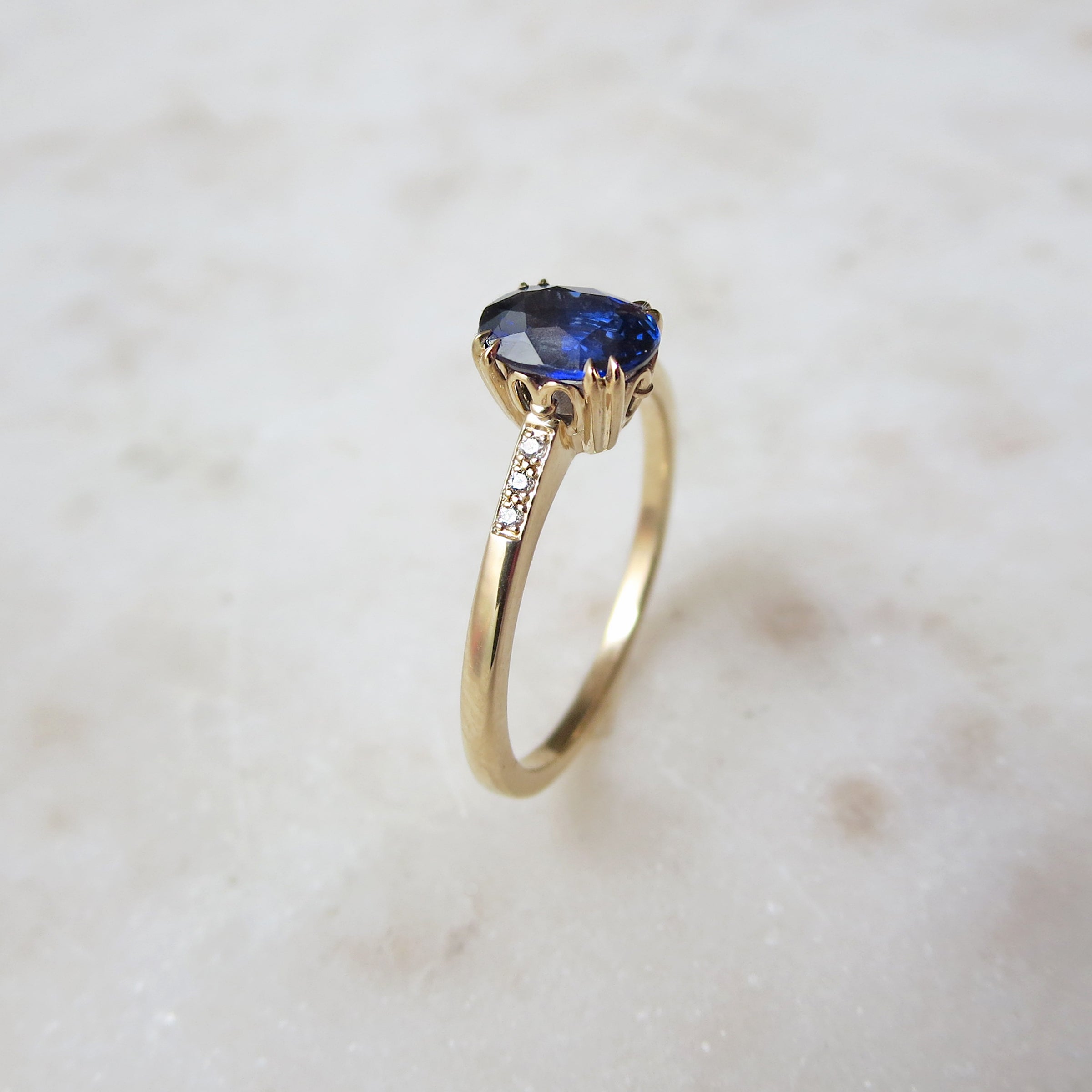 Angled view of a solitaire ring with an oval blue sapphire stone with white diamond pave on either side set in yellow gold on a grey background.