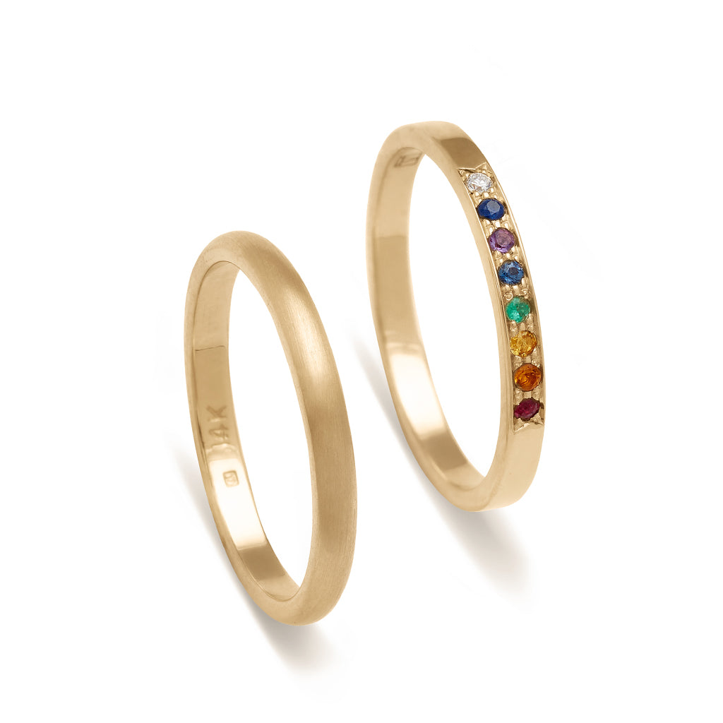A 2mm classic round band with a brushed finish set in yellow gold next to a 1.5mm band with rainbow gemstones set in yellow gold.