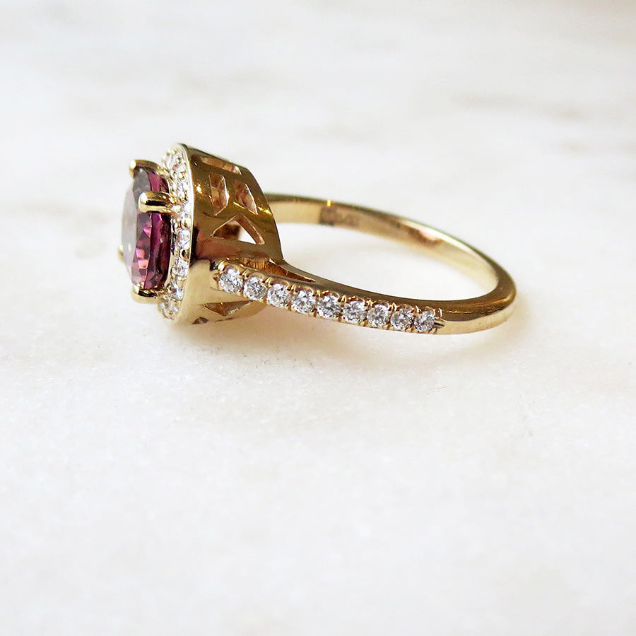 Side view of an engagement ring with an oval shape pink tourmaline center stone surrounded by a white diamond pave halo set in yellow gold with white diamond pave around the band on a grey background.