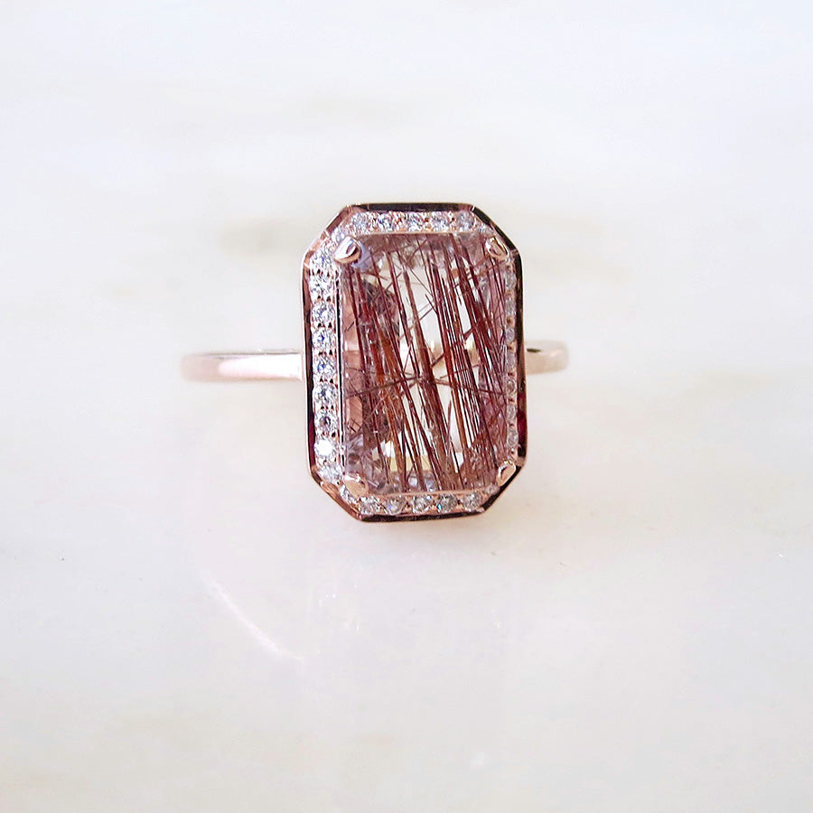 An engagement ring with an emerald shape copper rutilated quartz center stone surrounded by a white diamond pave halo set in rose gold on a grey background.