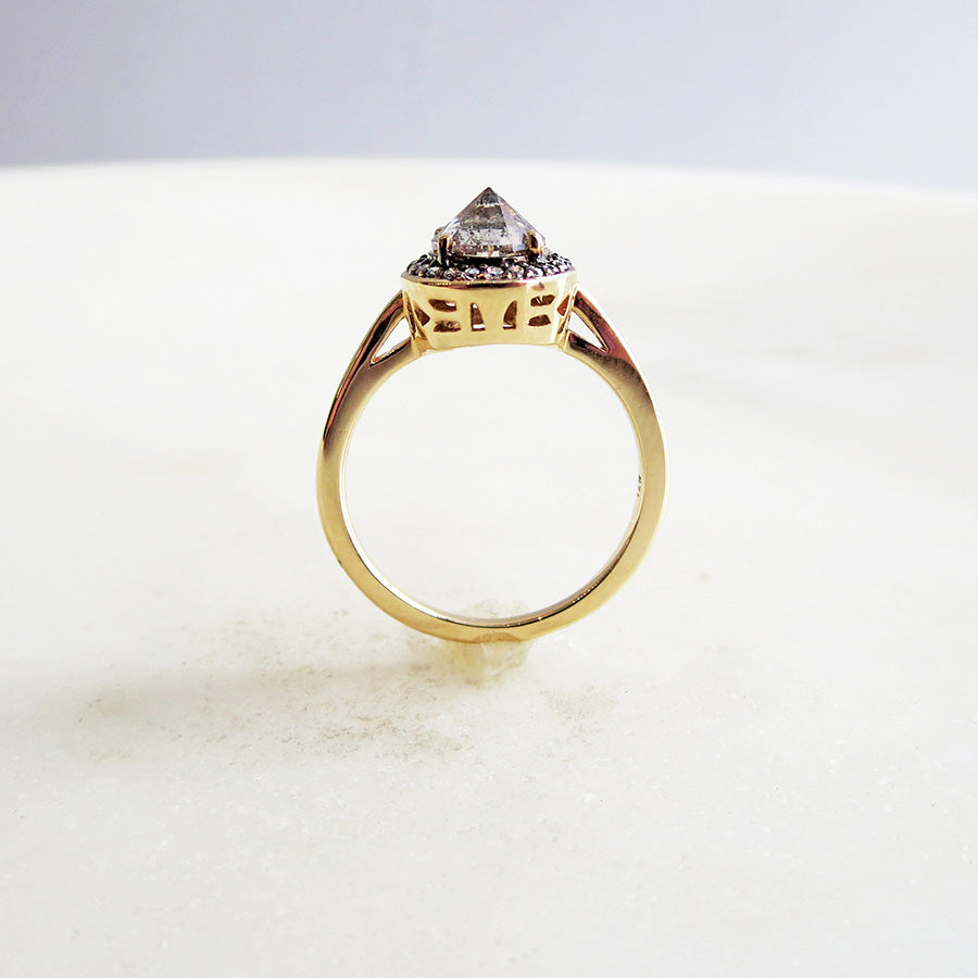 Side view of an engagement ring with a round shape inverted grey diamond center stone surrounded by a white diamond pave halo with black rhodium set in yellow gold on a grey background.