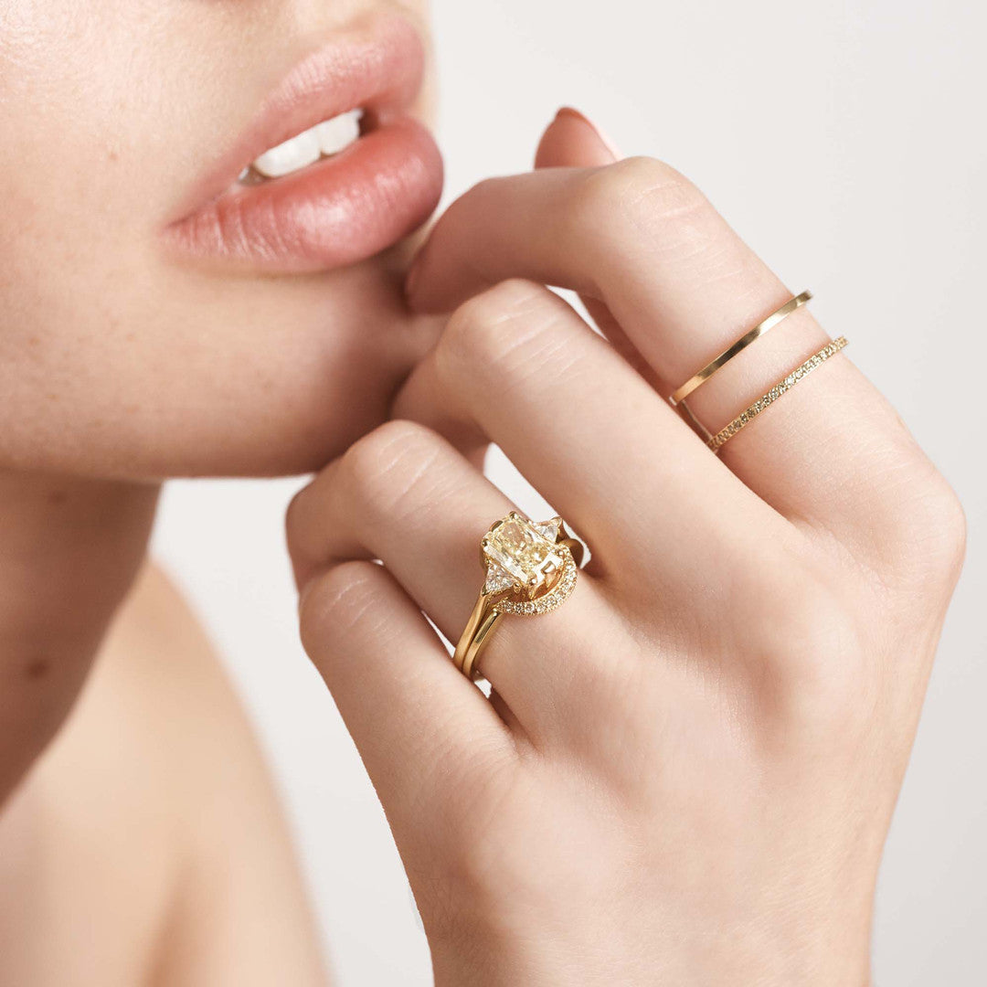 A three stone engagement ring with an emerald shape yellow diamond center stone with two white diamond trillion side stones set in yellow gold above a white diamond curved band on a light-complexion ring finger.