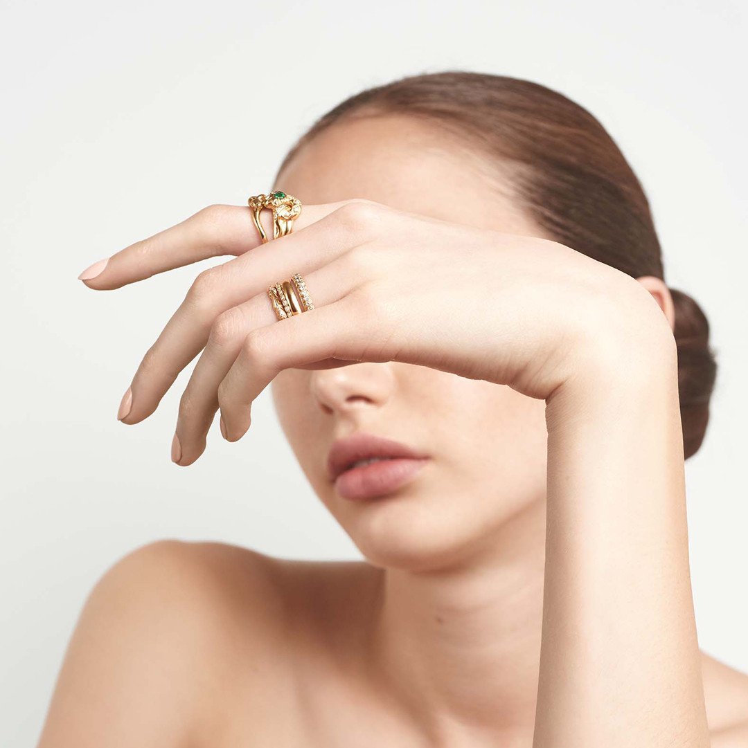 A serpent band with a green emerald center stone and white diamond accent stones around the head of the snake set in yellow gold on a lighter-complextion hand paired with another serpent band., White Gold, Rose Gold, Yellow Gold