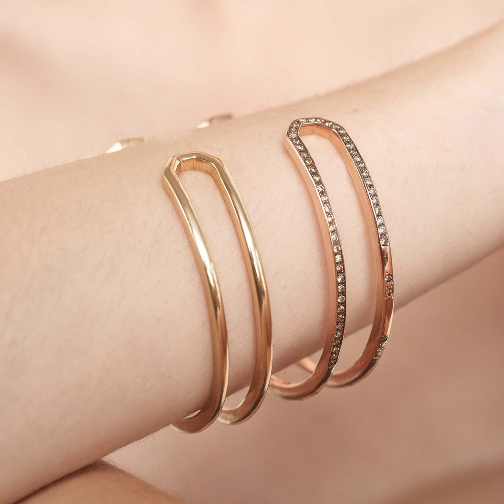 A doubled banded cuff bracelet set in yellow gold stacked above a diamond dusted version of the bracelet on a light-complextion model's wrist.