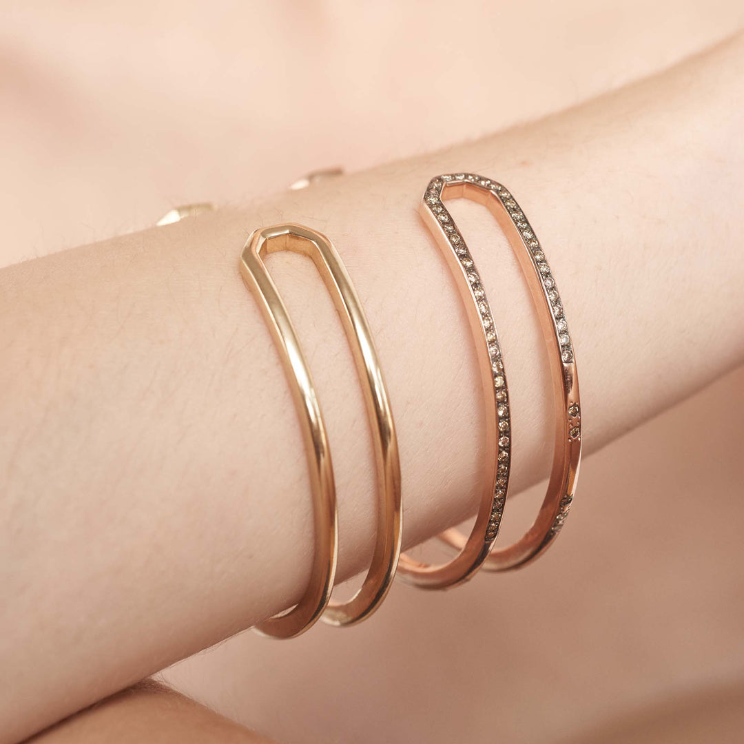 A doubled banded cuff bracelet set in yellow gold stacked above a diamond dusted version of the bracelet on a light-complextion model's wrist.