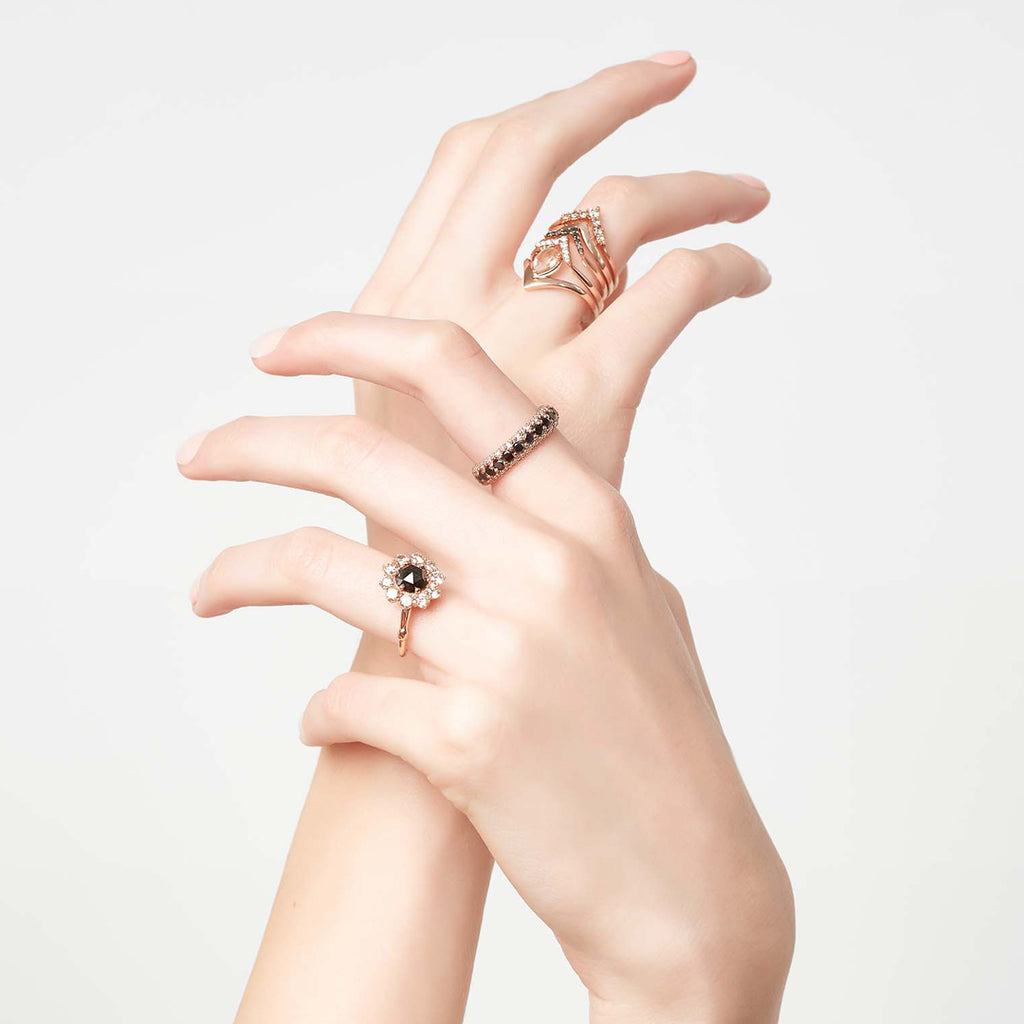 A ring with a rose cut black diamond center stone surrounded by round champagne diamonds set in rose gold on the ring finger of a light-complextion hand with chevron shaped bands stacked with a pear shaped orbit ring.