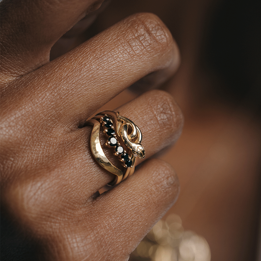 A serpent band with white diamonds for eyes set in yellow gold stacked above a black diamond curve band and a hammered solid gold curve band on a dark-complexion ring finger.