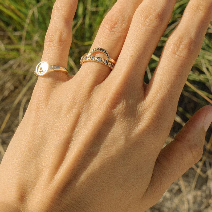 A 3mm straight band with white diamonds set in eternity around the band set in yellow gold stacked beneath a crescent shaped band on a light-complextion hand. On her pinky finger is an "A" monogrammed signet ring.