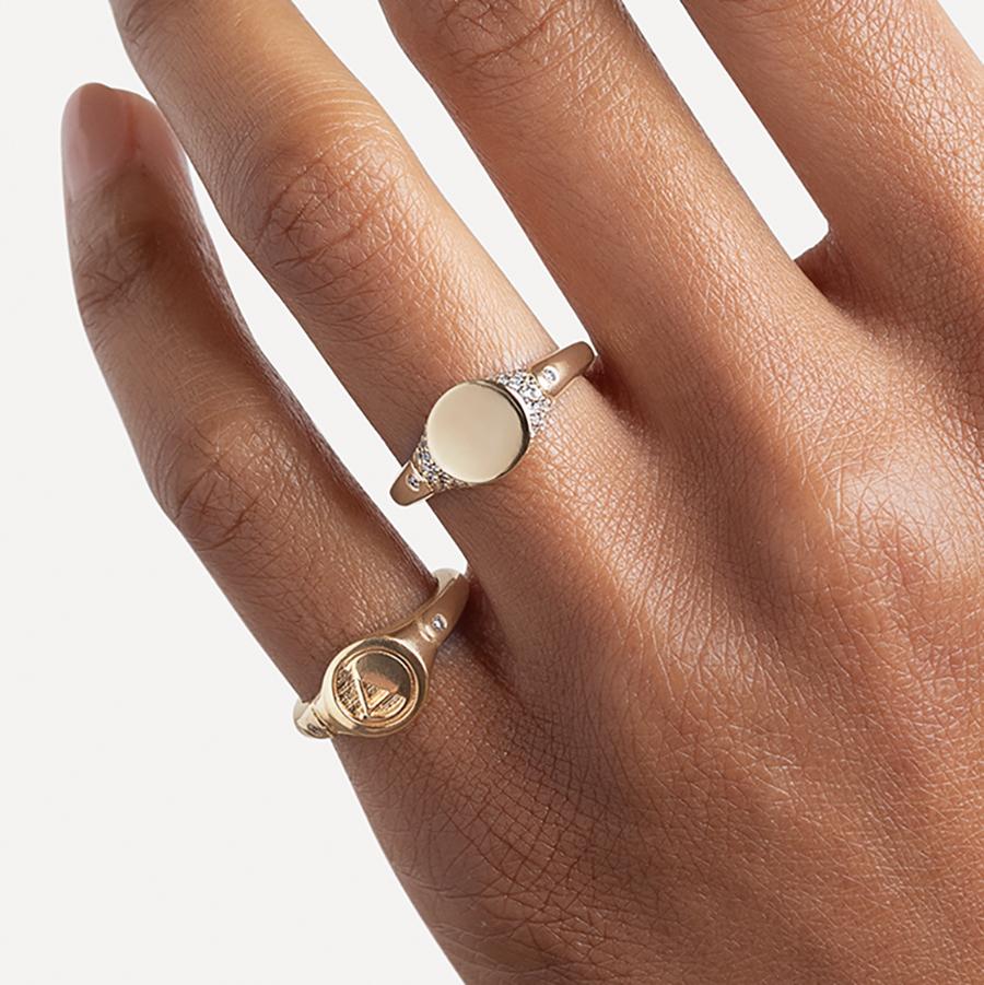 A solid gold signet ring with two white diamond accents on either side and a pave set around the top of the ring and set in yellow gold on a medium-complextion ring finger and a monogrammed signet ring with an "A" on the pinky finger.