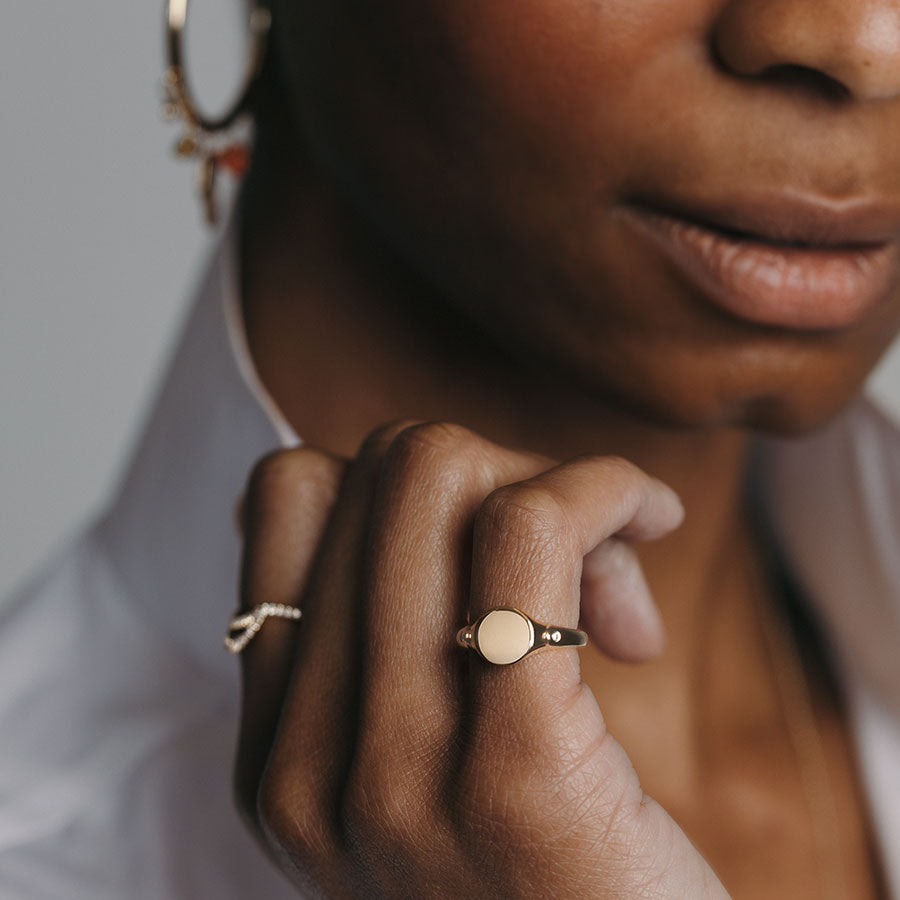A solid gold signet ring with two white diamond accents on either side and set in yellow gold on a darker-complextion pinky ring with a diamond accent band on her pointer finger.