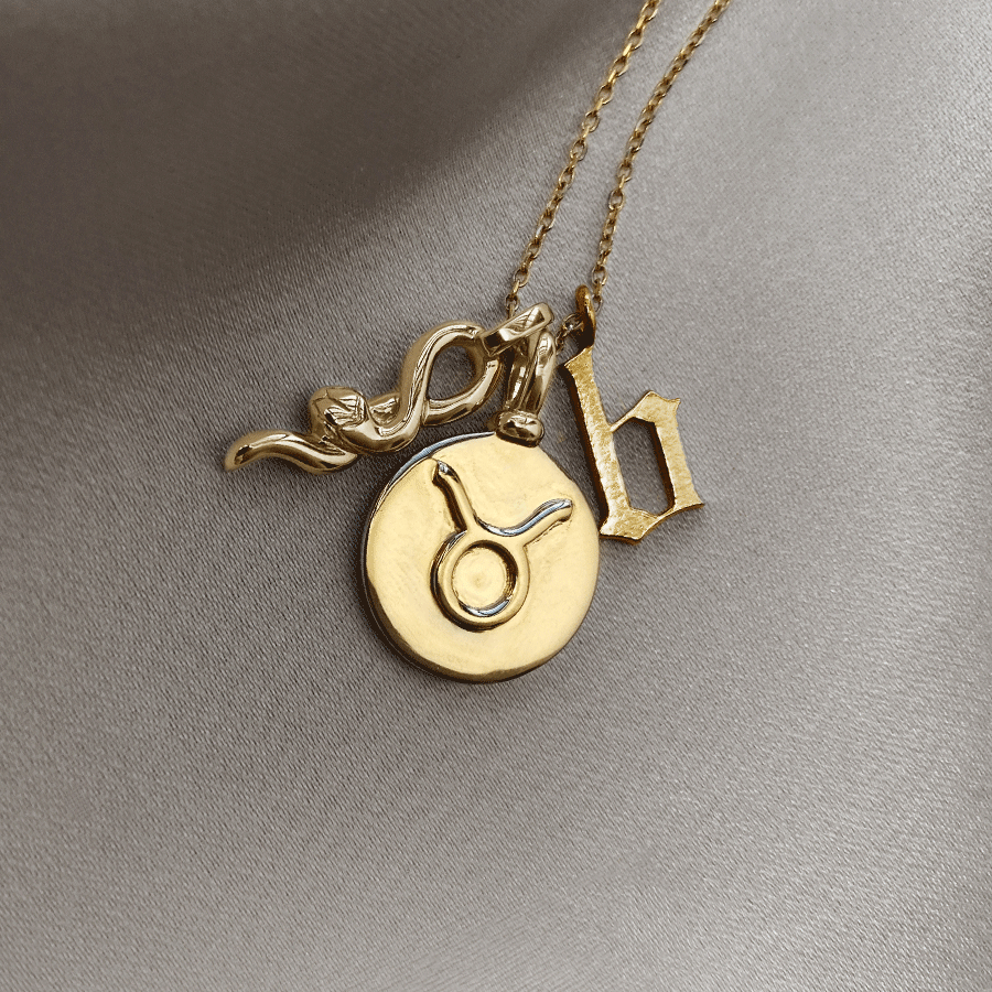 A 14mm charm with the Taurus zodiac sign set in yellow gold on a yellow gold chain with a medium serpent charm in yellow gold and an initial charm on grey fabric.