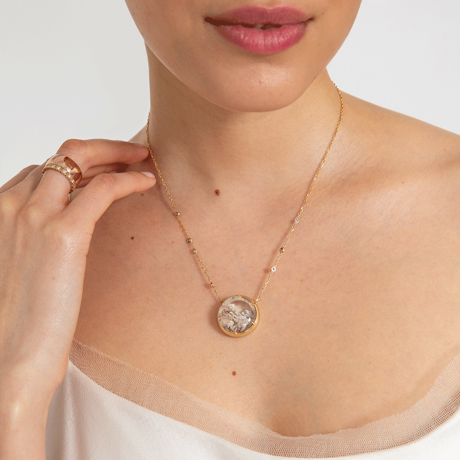 A quartz charm with a desert like scene in a bezel setting with claw prongs set in yellow gold on a yellow gold chain with grey diamonds on it on the neck of a light-complexion model.