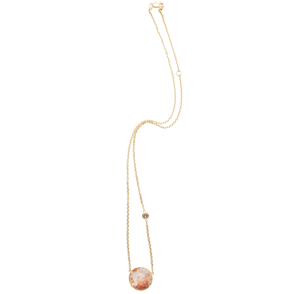 Zoomed out view of a round quartz charm with a desert like scene in a bezel setting with claw prongs set in yellow gold on a yellow gold chain with a champagne diamond on it.