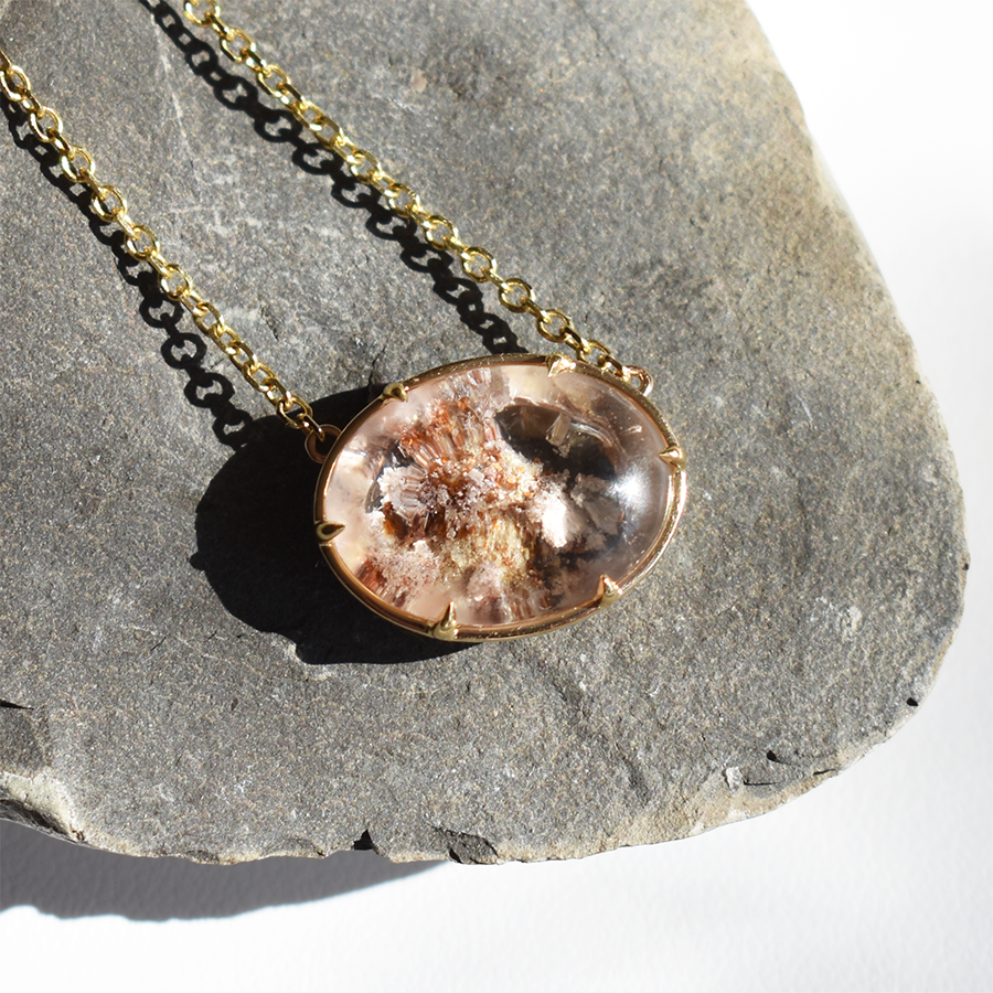 An oval quartz charm with a desert like scene in a bezel setting with claw prongs set in yellow gold on a yellow gold chain on a grey rock.